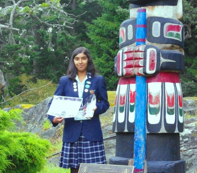 A student stands in front of a colourful totem pole with a certificate